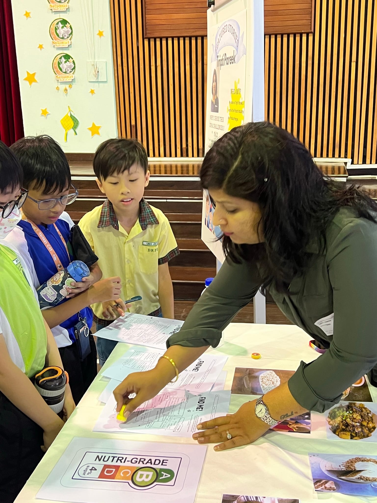 A woman points at papers on a table with a Nutri-Grade chart while three young students watch attentively.